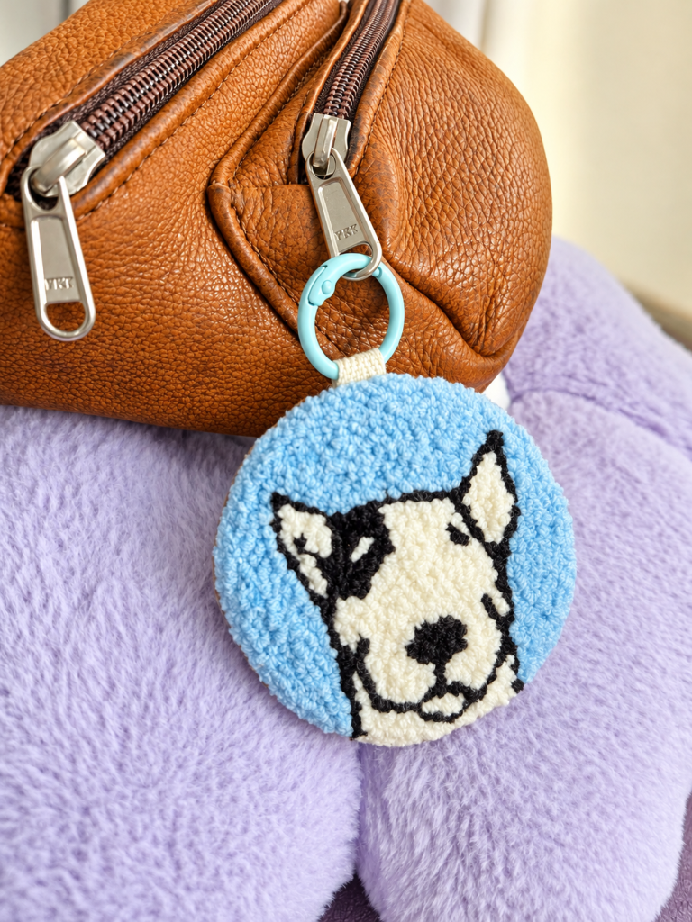 Handmade Rambo punch needle keyring and bag charm with a light blue background, black and white dog design and genuine leather backing, attached to a brown leather bag.