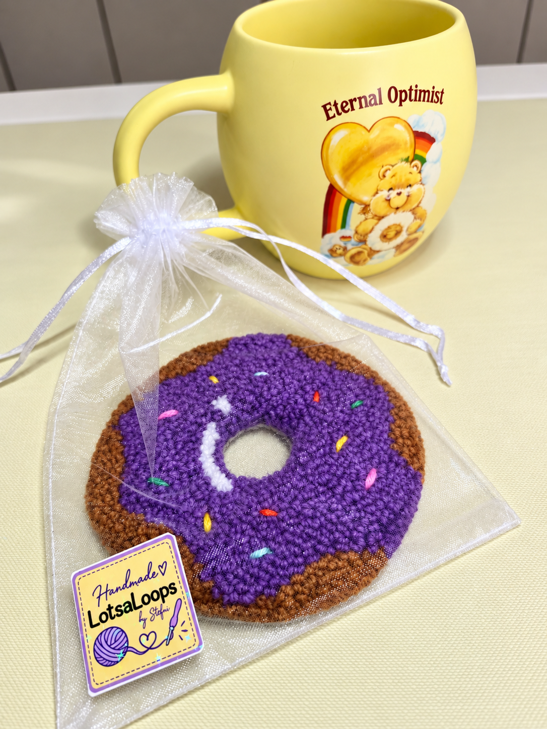 Purple donut punch needle mug rug in a white organza gift bag with LotsaLoops sticker, displayed beside a yellow mug.