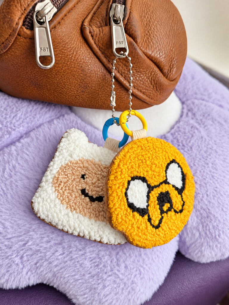 Handmade Finn and Jake punch needle duo bag charms with genuine leather backing, attached to a brown leather bag.