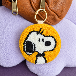 Handmade mustard-yellow Snoopy punch needle 2-in-1 keyring and bag charm with genuine leather backing, attached to a brown leather bag.