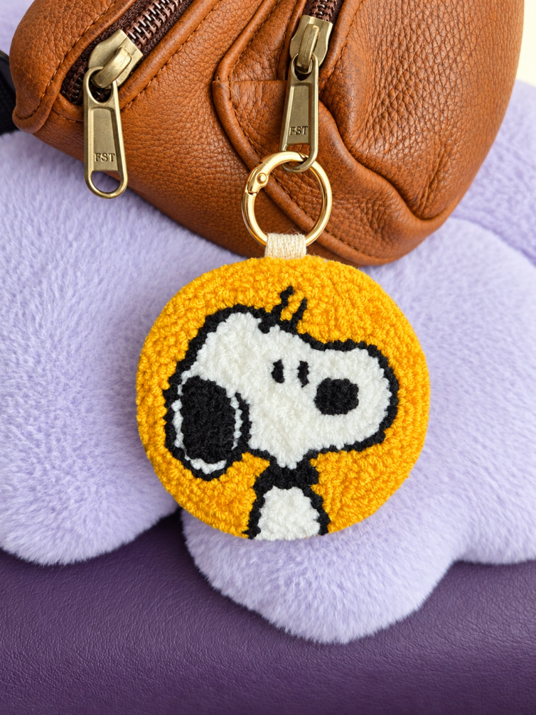 Handmade mustard-yellow Snoopy punch needle 2-in-1 keyring and bag charm with genuine leather backing, attached to a brown leather bag.